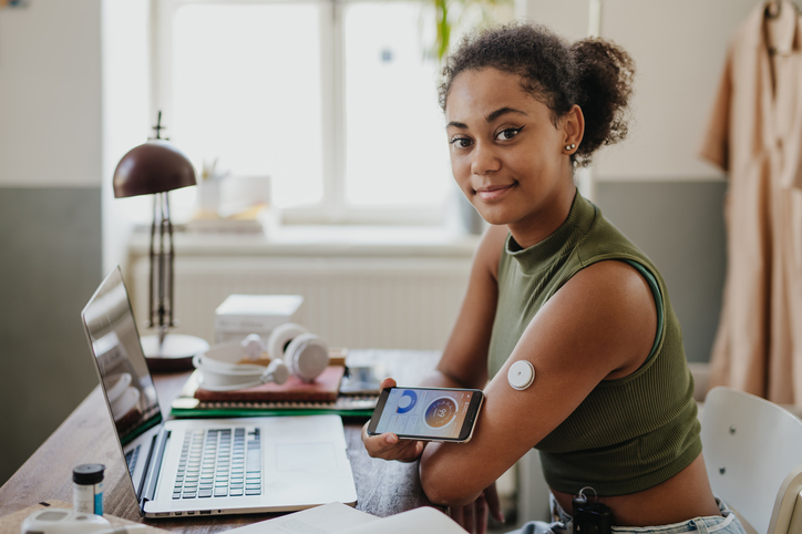 Teenage girl connecting smartphone to continuous glucose monitor, checking blood glucose during day. Student doing routine blood check while studying.